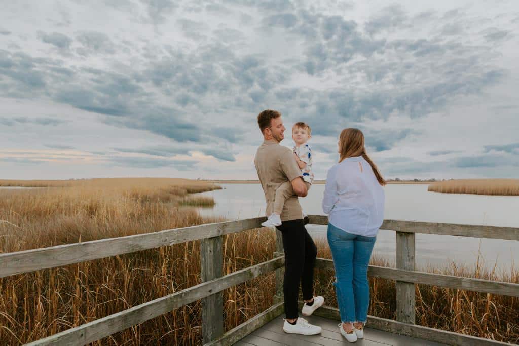 A family stands on a wooden dock by a calm marsh, admiring the scenery with a child held by a man and a woman beside him.