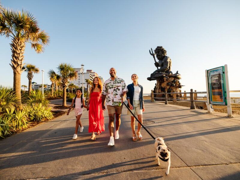 A family with two adults and two children walk along a palm-lined boardwalk, a dog on a leash, with the King Neptune statue in the background.