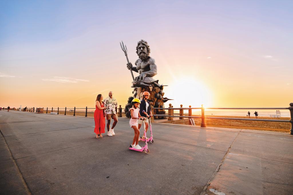 A group of people enjoying a sunset at a beach boardwalk with the King Neptune statue in the background.
