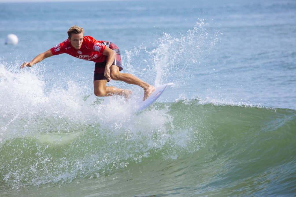 A professional surfer performs a dynamic maneuver on a green wave, creating splashes against a calm ocean backdrop.
