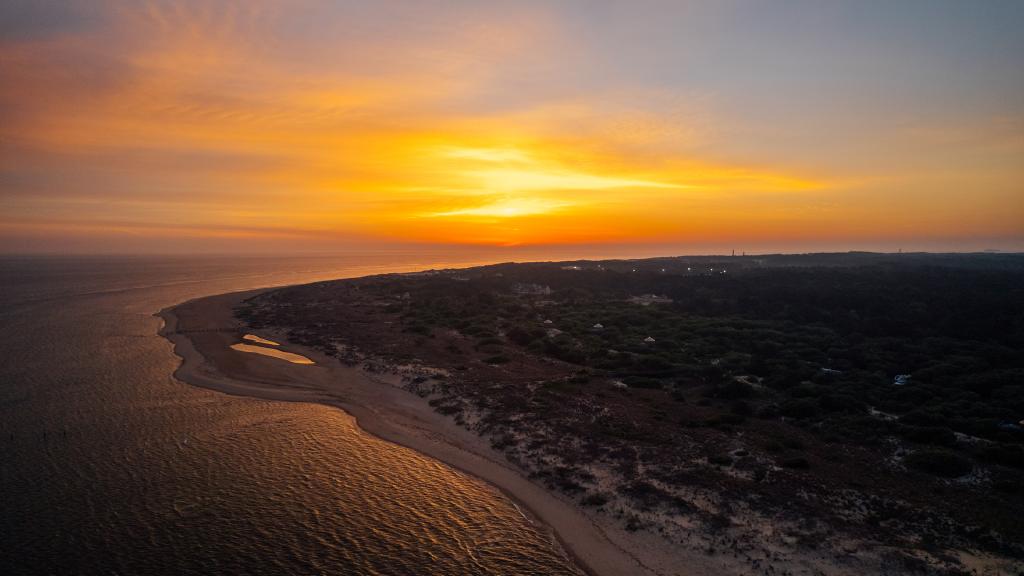 Aerial view of a serene beach at sunset, showcasing golden skies reflecting on calm waters and lush greenery in the background.