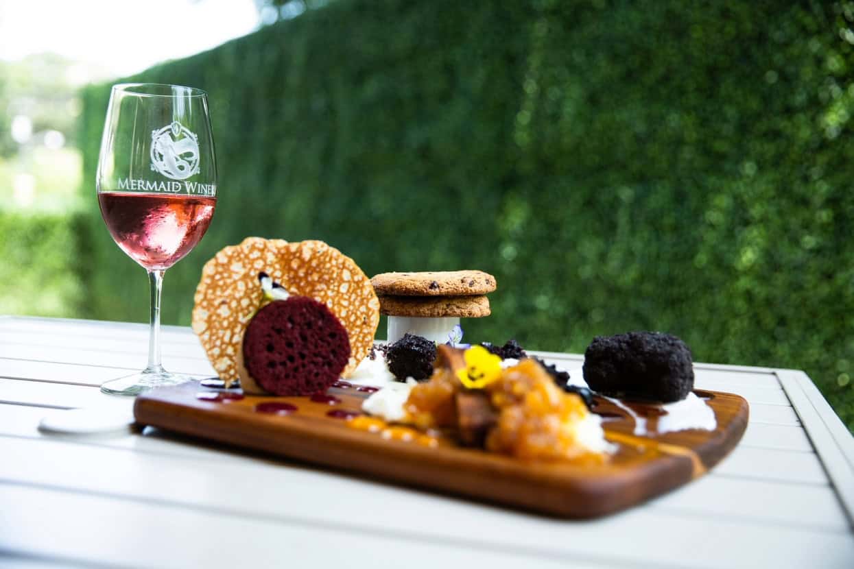 A glass of rosé wine beside a wooden platter featuring assorted desserts, including cookies, chocolate cake, and decorative garnishes.
