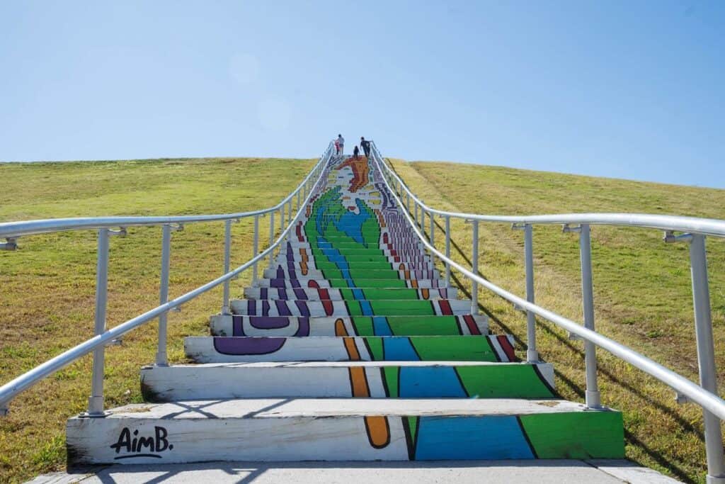 Colorful mural painted on stairs leading up a slope, with people walking towards the top under a clear blue sky.