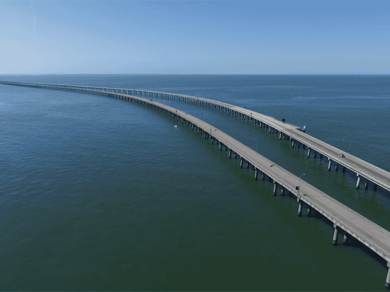 Aerial view of a curving bridge over calm blue waters under a clear sky.