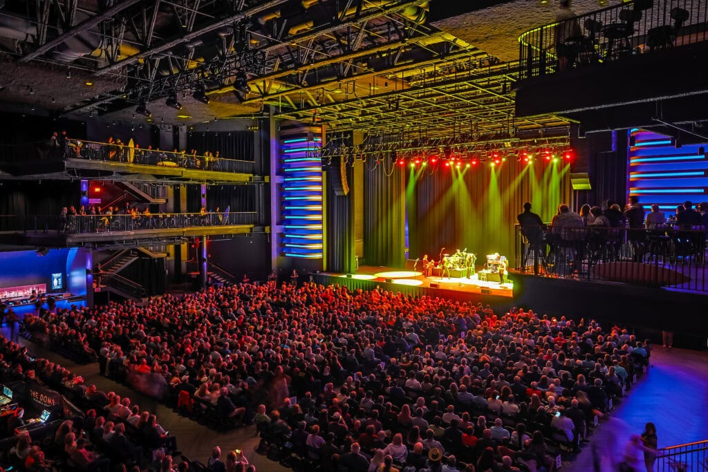 A large audience enjoys a live performance on stage, illuminated by colorful lights in a modern concert venue.