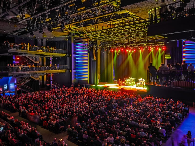 A large audience enjoys a live performance on stage, illuminated by colorful lights in a modern concert venue.