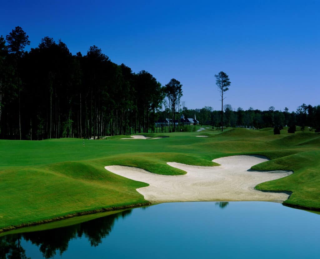 A scenic view of a well-maintained golf course featuring lush green grass, sand traps, and a tranquil pond under a blue sky.