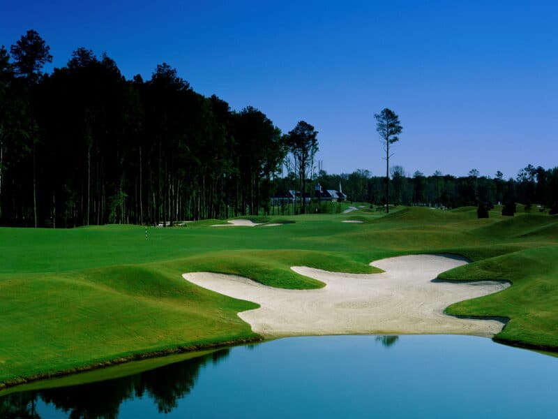 A scenic view of a well-maintained golf course featuring lush green grass, sand traps, and a tranquil pond under a blue sky.