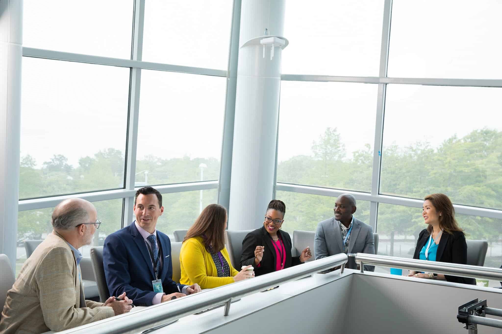 Business professionals engaged in discussion at a modern meeting space with large windows and green surroundings.