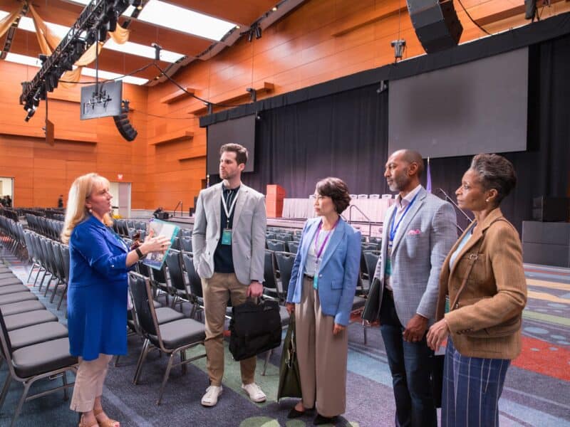 A professional group of four adults discusses details in a spacious conference hall with rows of chairs and a stage in the background.