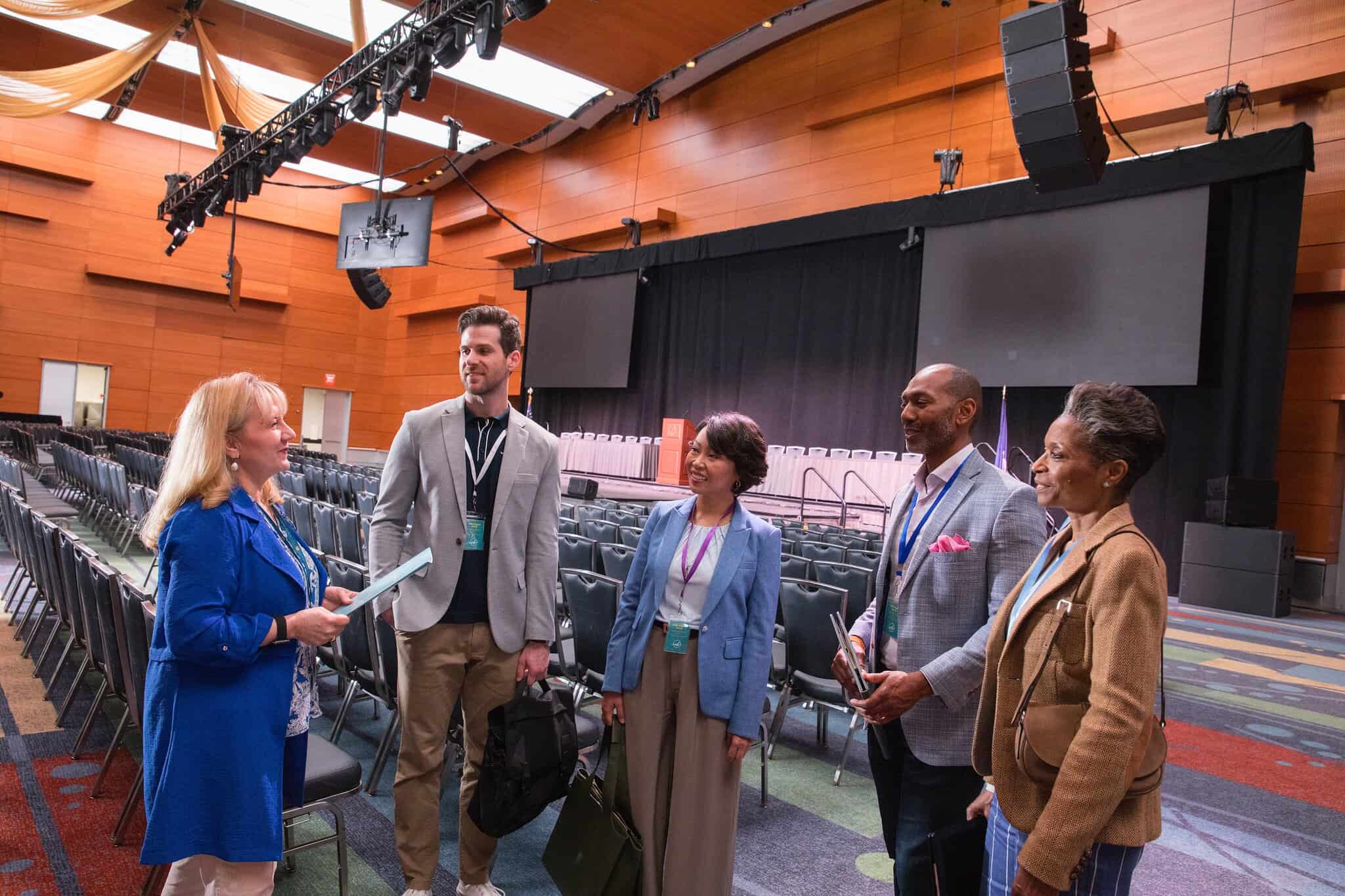 A group of five professionals conversing in a spacious conference hall with rows of chairs and a stage in the background.