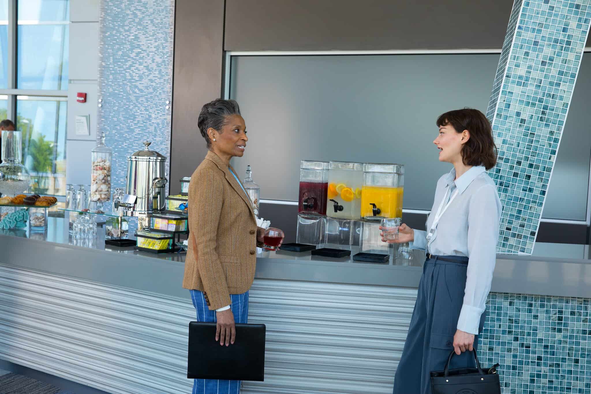 Two women engage in conversation by a modern refreshment station featuring drinks and snacks in a stylish setting.