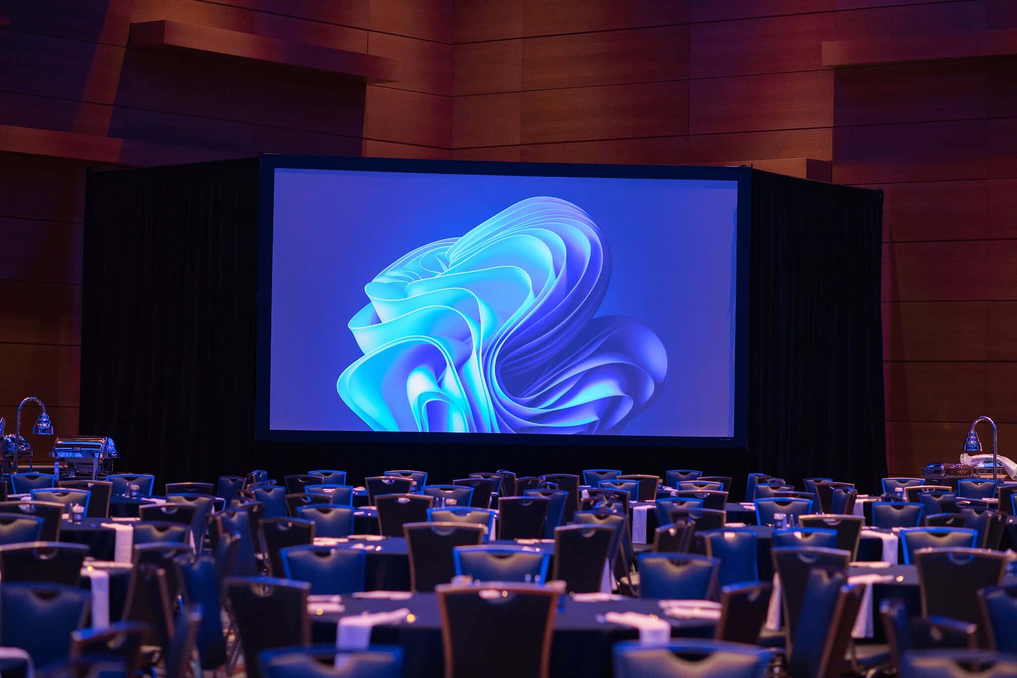 A large screen displaying a blue abstract design is set up in a banquet hall with numerous tables and chairs arranged throughout.