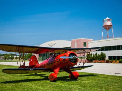 A bright red biplane parked on green grass in front of a vintage hangar under a clear blue sky.