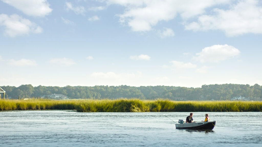 Two people fishing from a small boat on a calm river surrounded by lush green marshland under a blue sky with clouds.