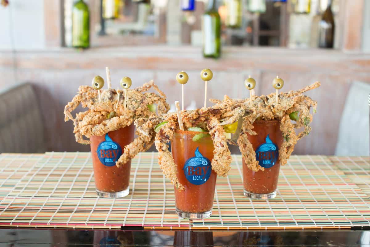 Three unique Bloody Mary cocktails topped with fried crab legs and olives, presented on a colorful striped table mat.