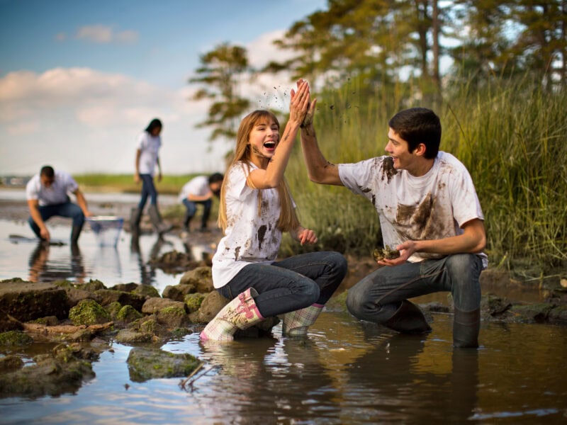 A group of young people joyfully participate in a muddy outdoor activity by the water, with two in focus giving a high five.