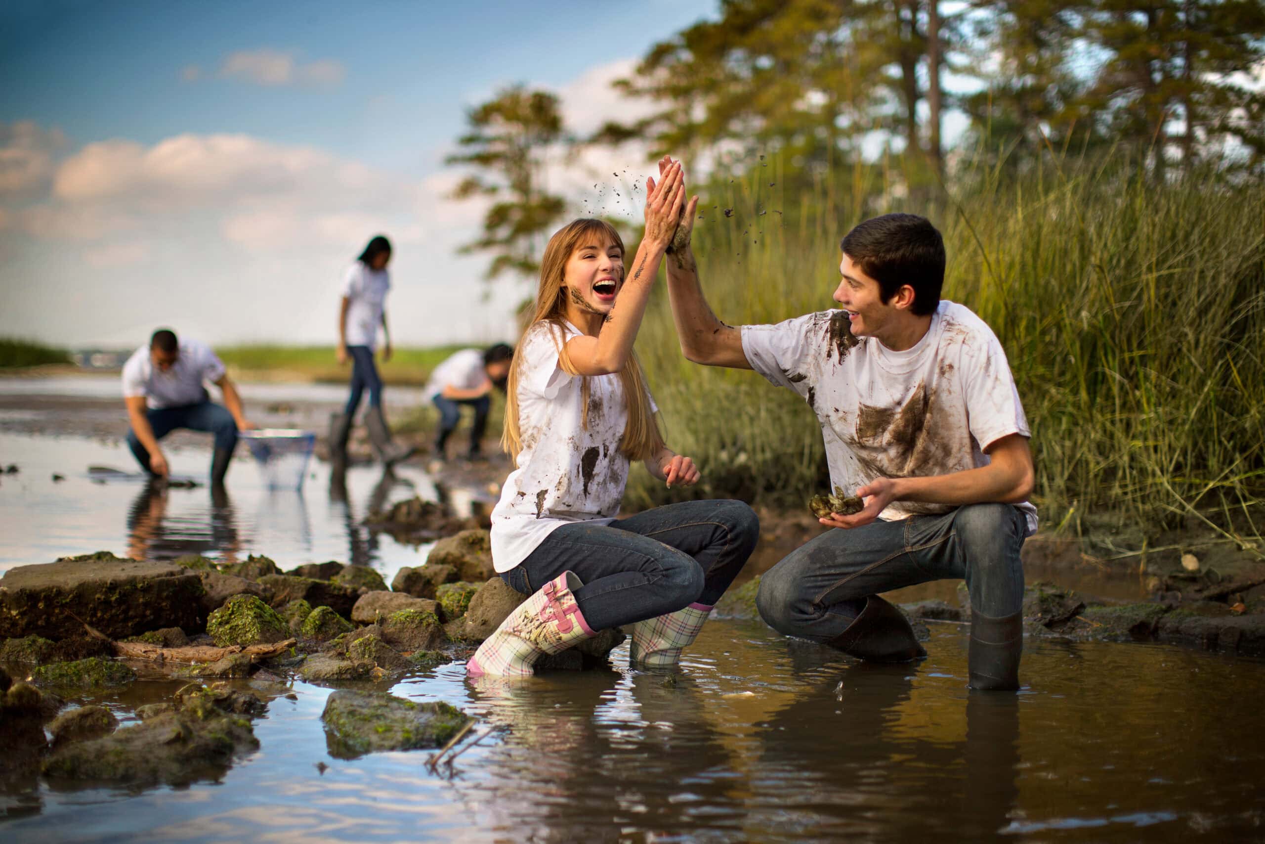 A group of young people joyfully participate in a muddy outdoor activity by the water, with two in focus giving a high five.