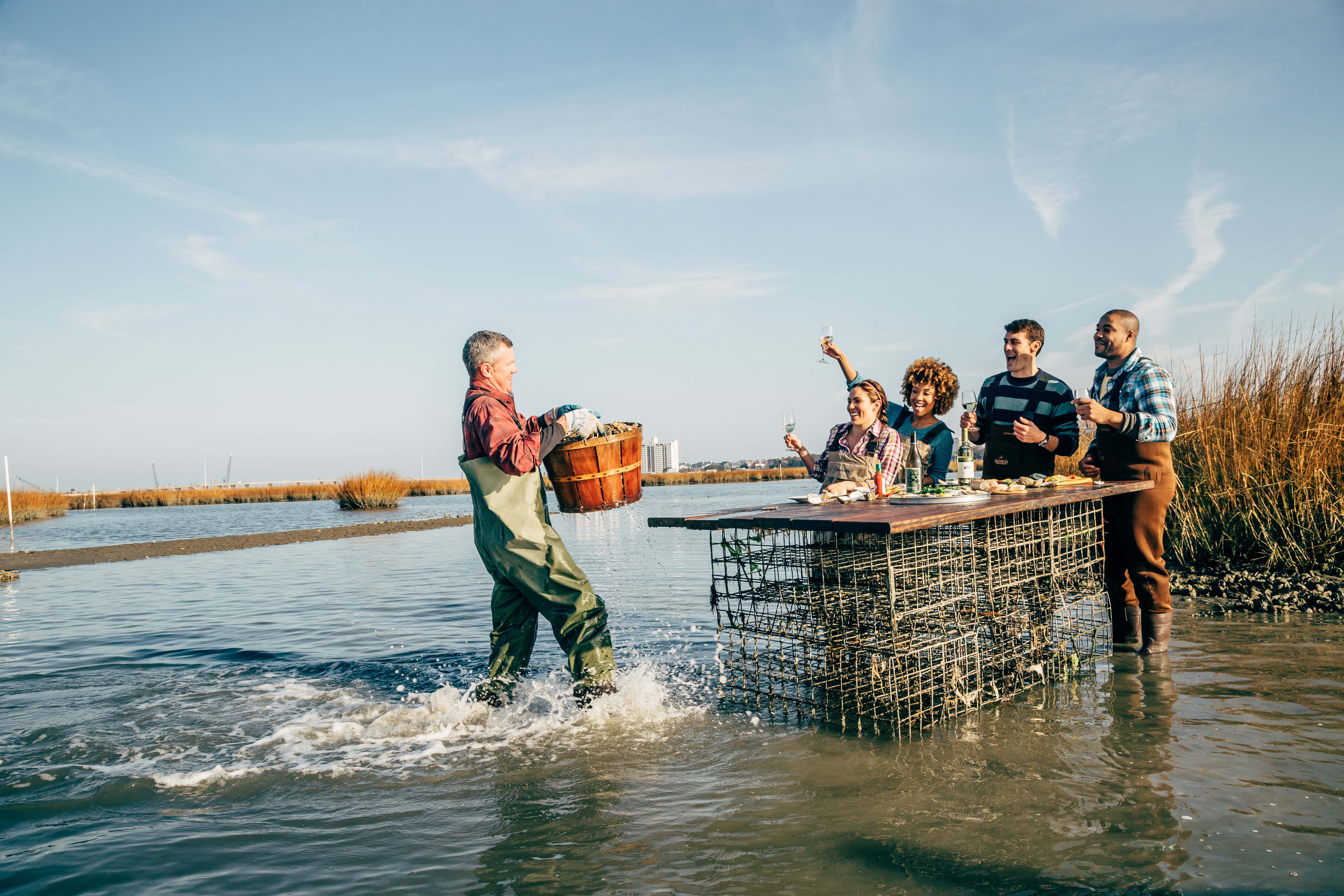 A group of five people celebrates along a waterfront with drinks while a man carries a basket of seafood at Pleasure House Oyster Farm.