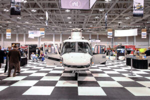 A helicopter on display at an exhibition, surrounded by attendees and featuring a checkered flooring design.