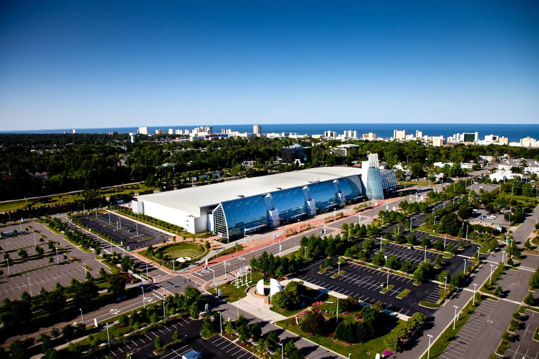 Aerial view of a large convention center surrounded by greenery and a parking lot, with a city skyline and lake in the background.