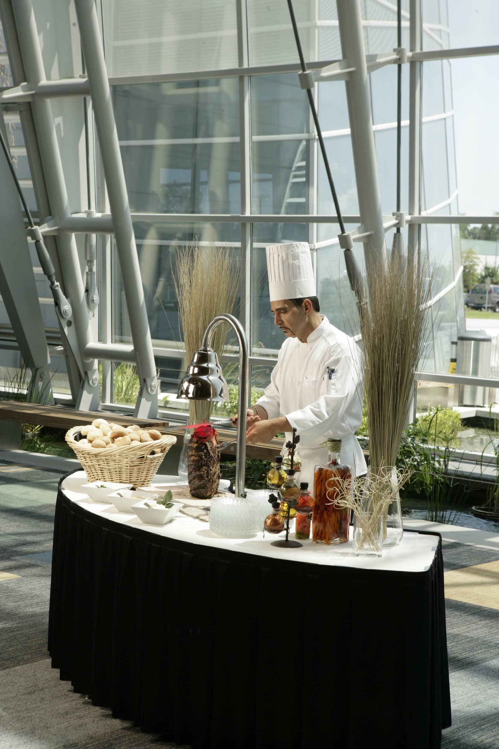 A chef in a white uniform and hat prepares food at a stylish catering table featuring bread rolls and various condiments, surrounded by large windows.