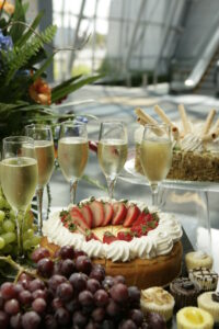 A festive dessert spread featuring a cheesecake topped with sliced strawberries, sparkling wine in fluted glasses, and an assortment of grapes and mini cupcakes.