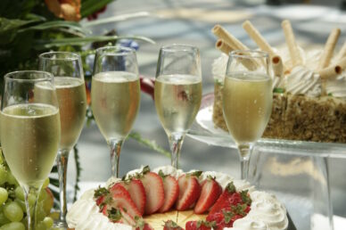 A festive dessert spread featuring a cheesecake topped with sliced strawberries, sparkling wine in fluted glasses, and an assortment of grapes and mini cupcakes.