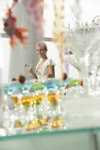 A cheerful woman in a white dress holds a colorful cocktail amidst a display of various drinks at a vibrant event.