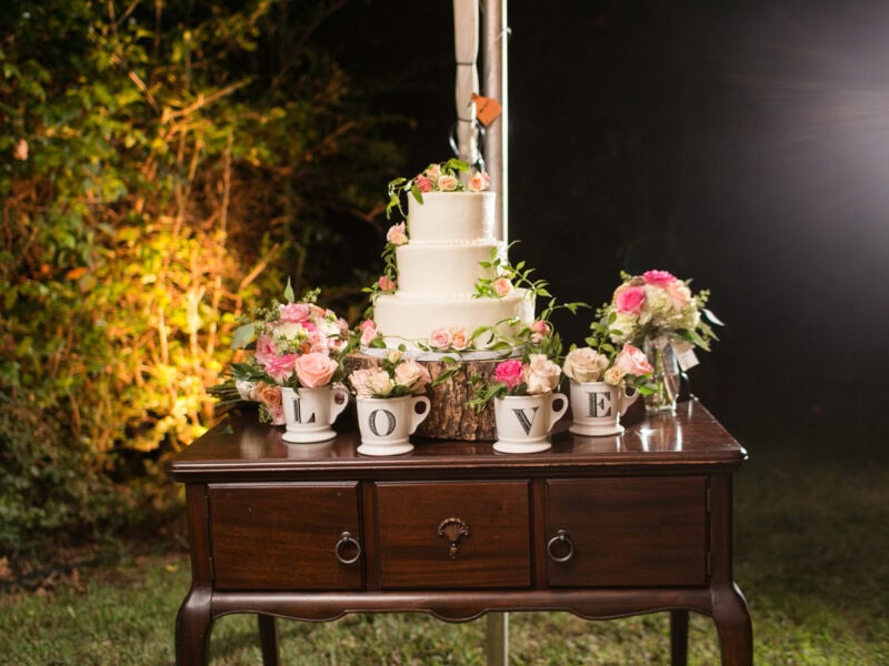 A beautifully arranged wedding cake with floral decorations, set on a wooden table adorned with flower bouquets and cups spelling "LOVE."