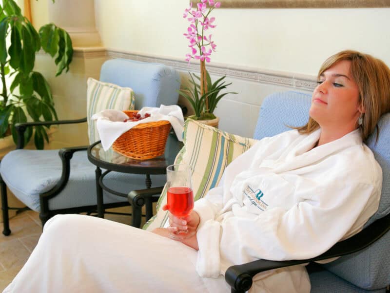 A woman in a white robe relaxes in a spa setting, holding a glass of red beverage, surrounded by plants and comfortable seating.