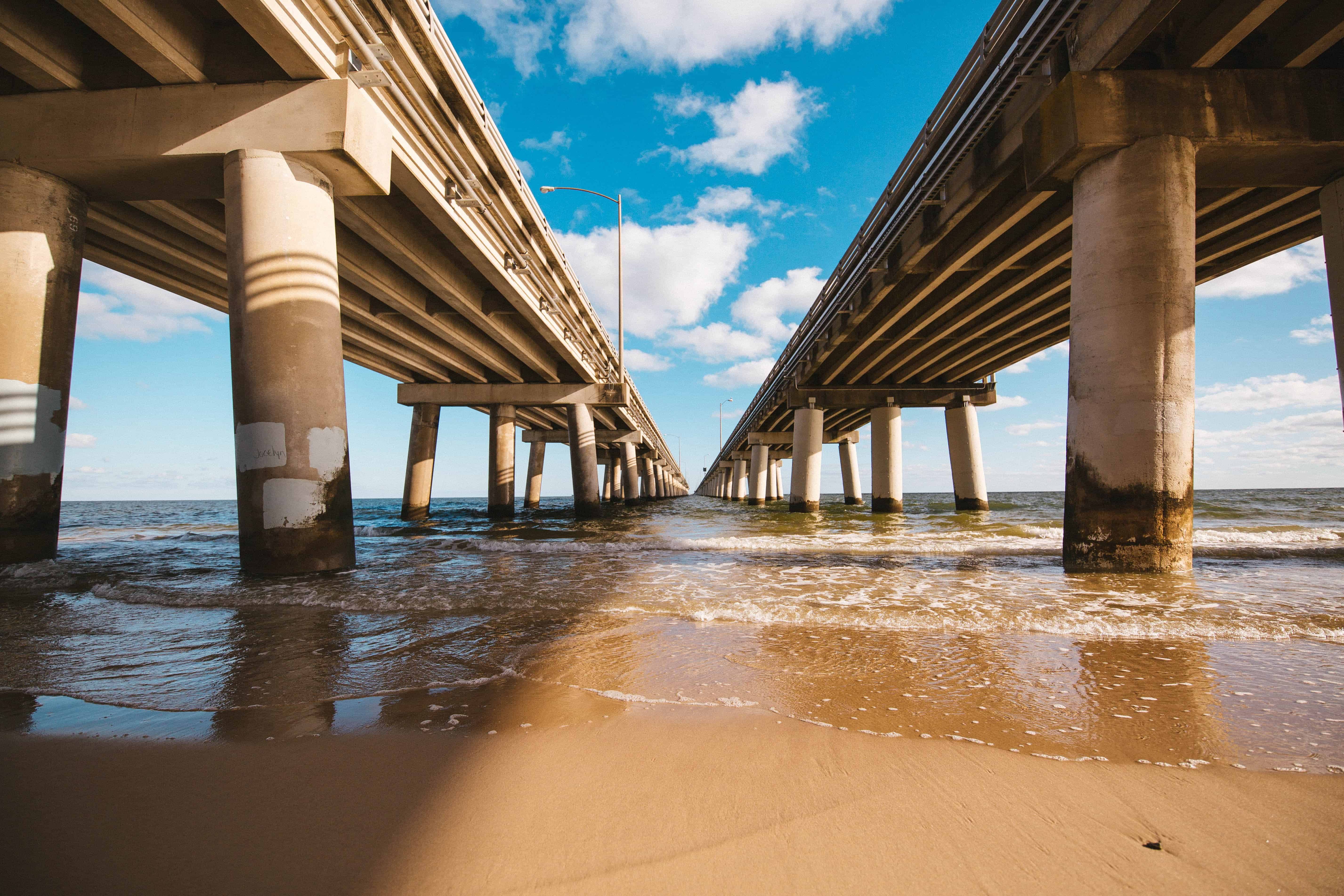 Underneath a highway bridge, with concrete pillars above sandy beach and gentle waves, under a bright blue sky with clouds.