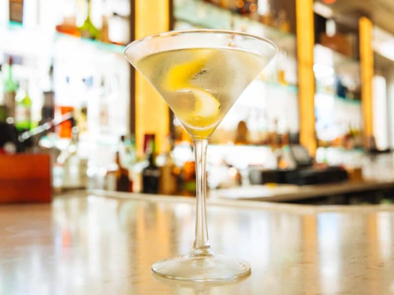 A close-up of a classic martini glass with a lemon twist, set against a well-stocked bar background.