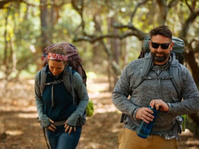 Two hikers, a man and a woman, are walking through a sunlit forest trail, each carrying backpacks and enjoying the outdoors.