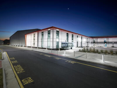 Modern building exterior at dusk, featuring glass panels, red accents, and illuminated pathways with 'No Parking' signs.