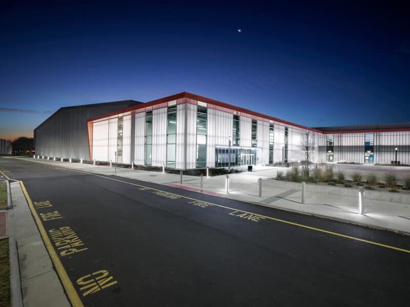 Modern building exterior at dusk, featuring glass panels, red accents, and illuminated pathways with 'No Parking' signs.