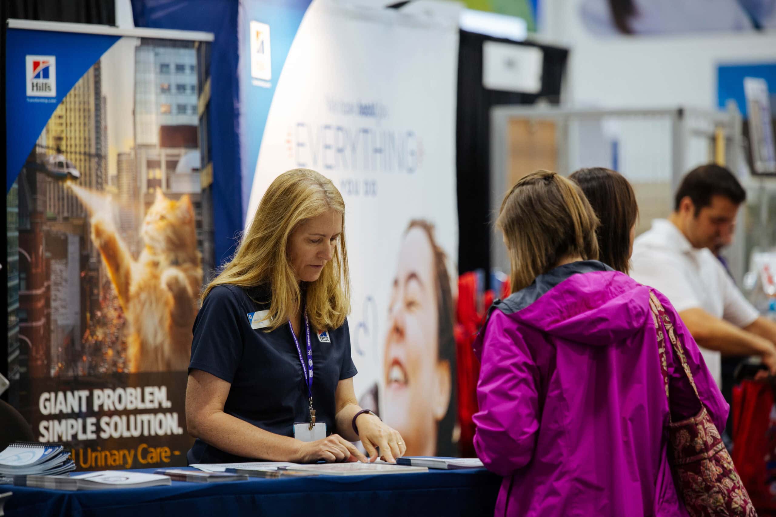 A woman at a booth is engaging with attendees, providing information about pet urinary care products at an event.
