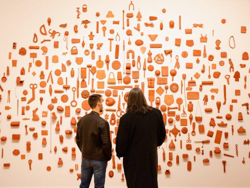 Two viewers observe a wall installation featuring an array of red ceramic objects, showcasing artistic reinterpretations of everyday items.