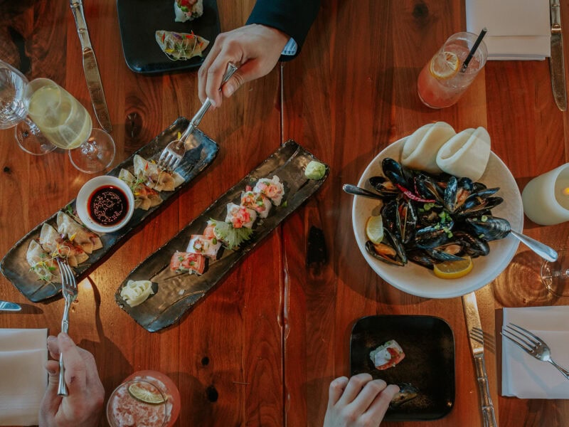 A top-down view of a dining table featuring sushi, dumplings, mussels, and drinks, with hands reaching for the dishes.