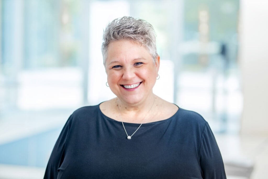 Smiling woman with short gray hair wearing a black blouse and necklace, standing in a bright, modern environment.