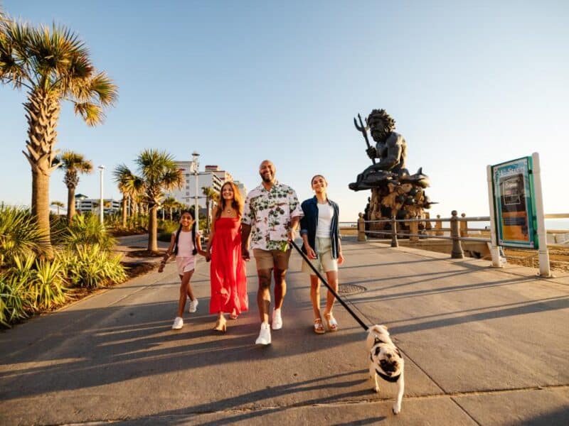 A family strolls along a palm-lined boardwalk, passing a large statue, with a dog on a leash and beachside buildings in the background.