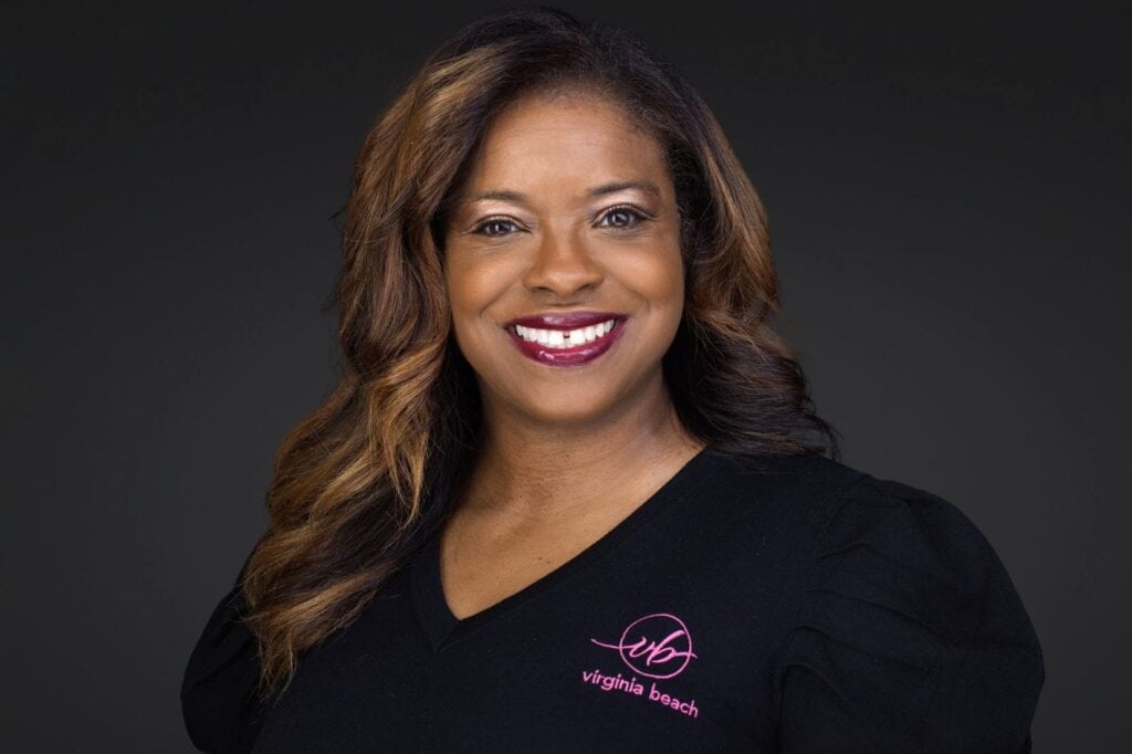 Smiling woman with long, wavy hair wearing a black top with "Virginia Beach" logo, set against a dark background.