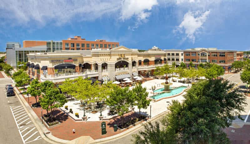 Aerial view of a vibrant outdoor shopping plaza featuring a fountain, greenery, and various shops under a blue sky.
