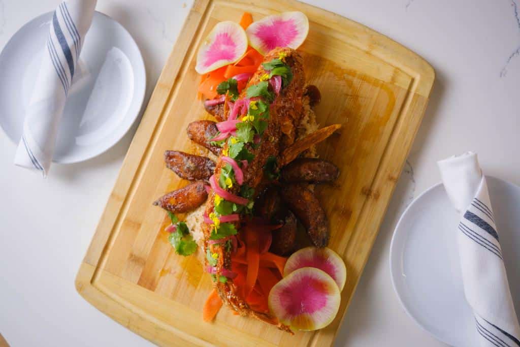 A beautifully arranged dish featuring a fried whole fish topped with vibrant garnishes like cilantro, pickled red onions, and colorful radish slices, served on a wooden board.