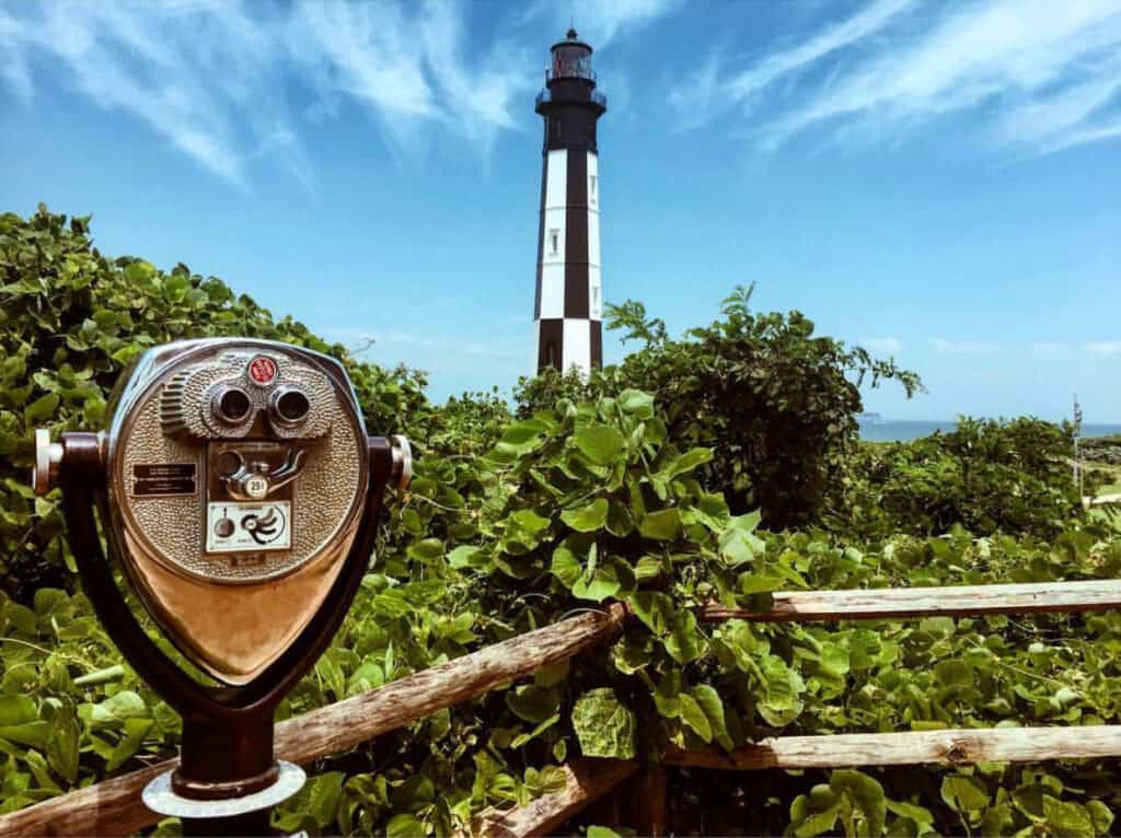 A vintage binocular viewer stands in the foreground with the new Cape Henry lighthouse and blue skies in the background, surrounded by greenery.