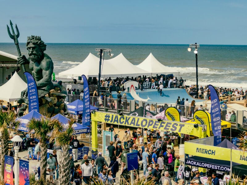 A vibrant beach festival scene featuring a large statue of Neptune, skateboarding activities, crowds, and colorful tents under a clear blue sky.