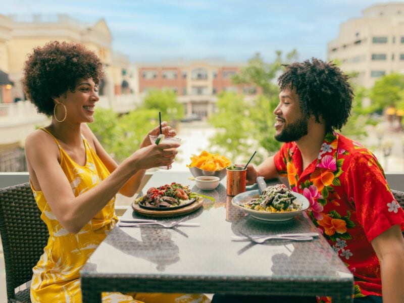 A couple enjoys a casual dining experience on a balcony at Cantina Laredo, sharing drinks and a meal, with vibrant dishes and a lively atmosphere with Town Center behind them.