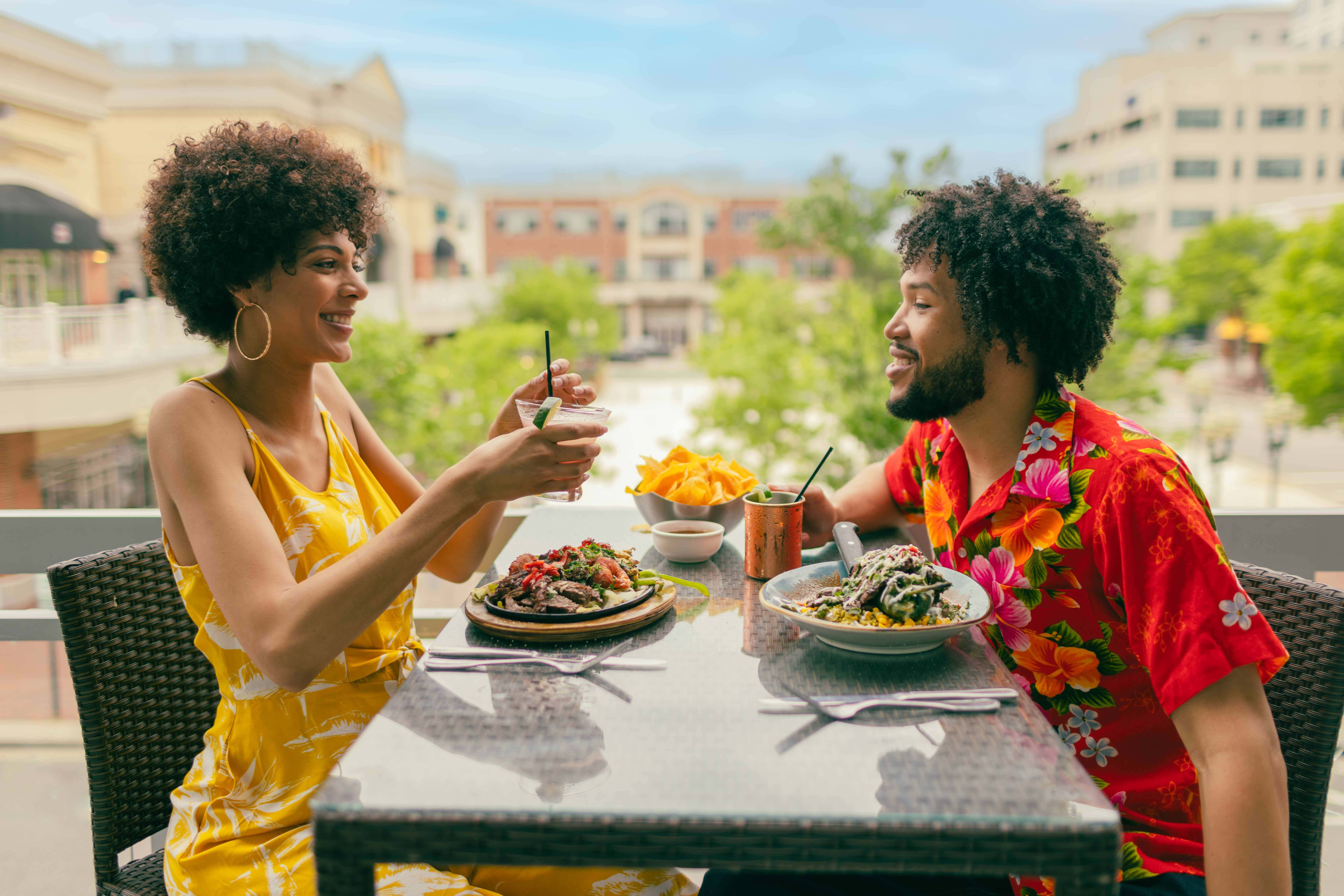 A couple enjoys a casual dining experience on a balcony at Cantina Laredo, sharing drinks and a meal, with vibrant dishes and a lively atmosphere with Town Center behind them.