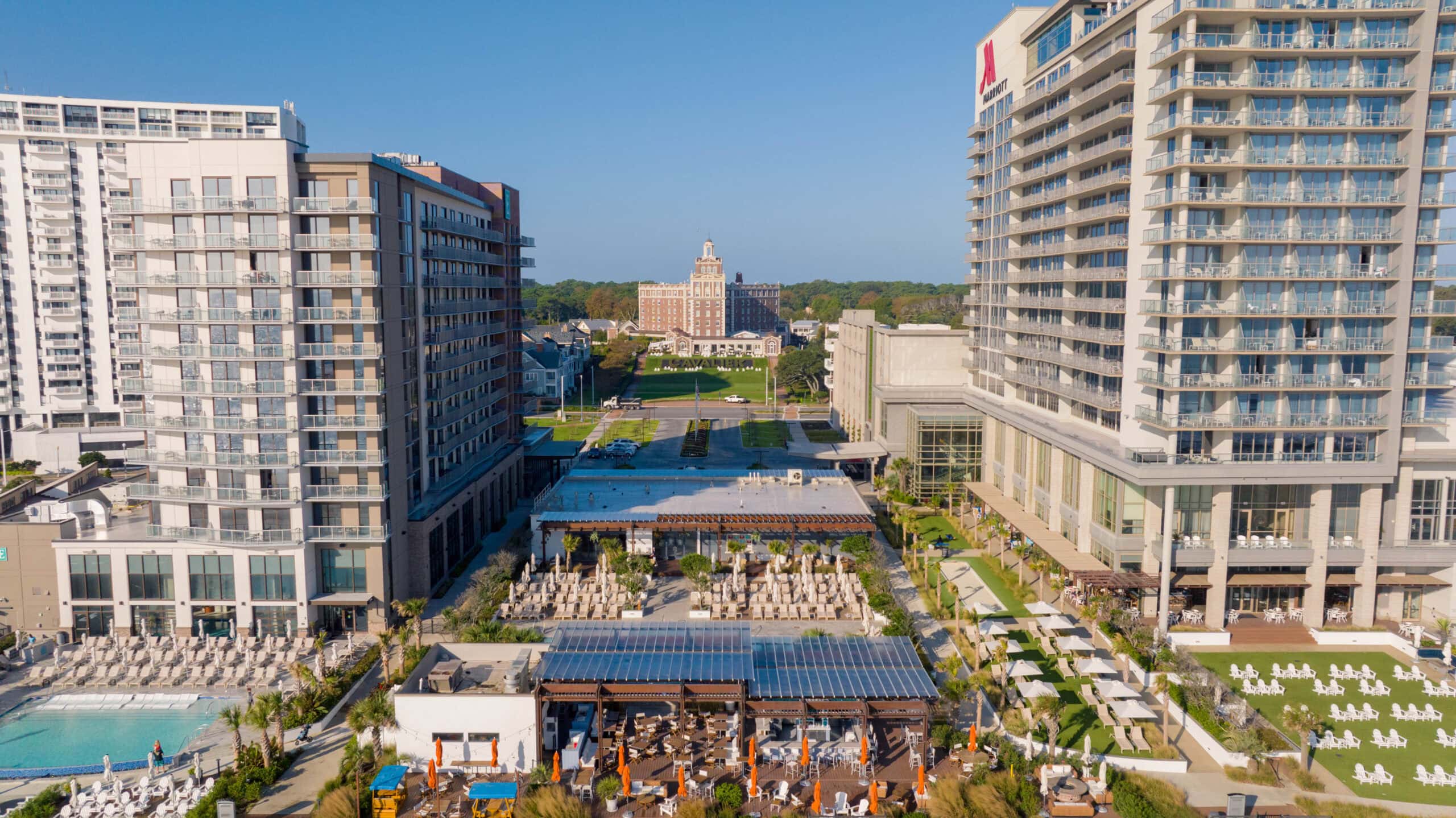 A panoramic view of a luxury hotel resort featuring multiple buildings, a pool area, and landscaped green spaces on a sunny day at the Virginia Beach Oceanfront.