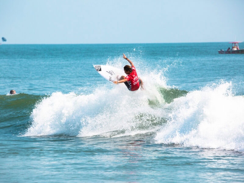 A surfer in a red jersey performs an aerial maneuver on a wave, with a boat visible in the background on a sunny day.